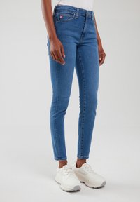 Inimene, kes kannab istuvaid siniseid teksapükse ja valgeid tossusid, seisab ühetaustal heleda tausta ees.