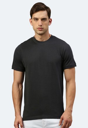 Junger Mann mit kurzen braunen Haaren, der ein schlichtes schwarzfarbenes T-Shirt mit kurzen Ärmeln und Rundhalsausschnitt vor einem hellgrauen Hintergrund trägt.