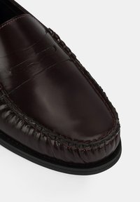 Bordeaux leren loafer met een gladde textuur, gestikte details en een zwarte rubberen zool. Heeft een decoratieve band over de bovenkant.