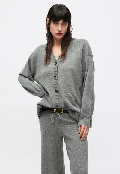 Sivý pletený kardigán s výstrihom do V, prednými gombíkmi a bočnými vreckami v kombinácii so zodpovedajúcimi rebrovanými nohavicami; mäkká textúra a pohodlný strih.
