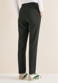 Donkergroene broek met een gestructureerde afwerking, elastische tailleband en twee achterzakken, gecombineerd met witte en groene sneakers.