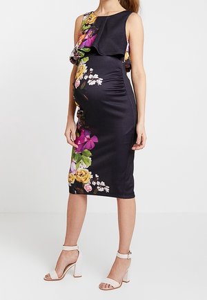 Schwangere Frau trägt ein eng anliegendes, ärmelloses, knielanges schwarzes Kleid mit buntem Blumenmuster und weiße Sandaletten mit Absatz.