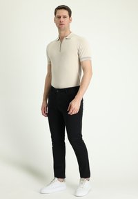 Polo beige de manga corta con cuello de cremallera, combinado con pantalones negros ajustados y zapatillas blancas, con una textura suave y un diseño minimalista.
