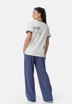 Tumehobusega naine, kes kannab valget T-särki, millel on kiri „CARDIO BUNNY“, ja siniseid laiade pükstega, seisab seljaga valge tausta ees.