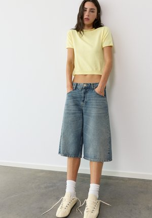 Frau mit langen dunklen Haaren, die ein gelbes kurzes T-Shirt, lockere knielange Jeans-Shorts, weiße Socken und hellbeige Sneakers trägt, steht an einer weißen Wand.