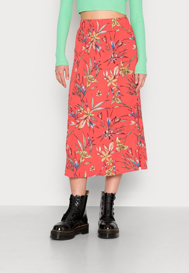 Jupe midi corail avec un motif floral en jaune, bleu et vert. Équipée d'une taille élastique et tombe de manière fluide. Associée à des bottines noires.