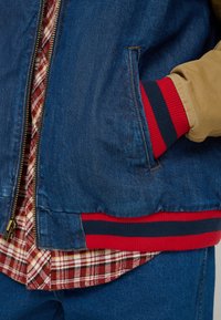 Denim jacket with two side pockets, a mustard lining, and red and navy striped ribbed cuffs layered over a red plaid shirt.