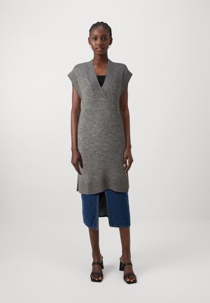 Robe tricotée grise avec un col en V et des manches courtes, présentant des bords côtelés et une fente sur le côté, superposée à une jupe en denim. Sandales à talons noires.