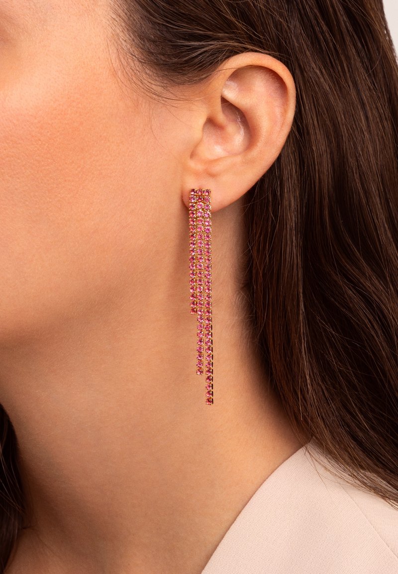 Oreille d’une femme avec une longue boucle d’oreille chandelier en strass rose, cheveux bruns et lisses, et vêtement beige clair visible au niveau du cou et de l’épaule.