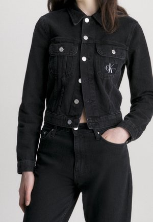 Woman wearing a fitted black denim jacket with silver buttons and matching black jeans, hand in pocket, cropped at waist.