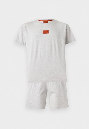 Homme debout pieds nus portant un short gris clair et un T-shirt assorti avec un petit logo rouge "HUGO" sur la poitrine, mains dans les poches.