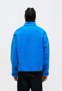 Niebieska polarowa kurtka z wysokim kołnierzem, teksturowaną powierzchnią i luźnym krojem. Posiada opadające ramiona i ściągacze przy mankietach.