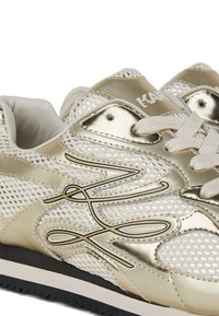 Baskets de sport métalliques dorées avec des panneaux en maille, des lacets crème, des accents texturés et une semelle en caoutchouc noir. Présente un logo sur la languette.