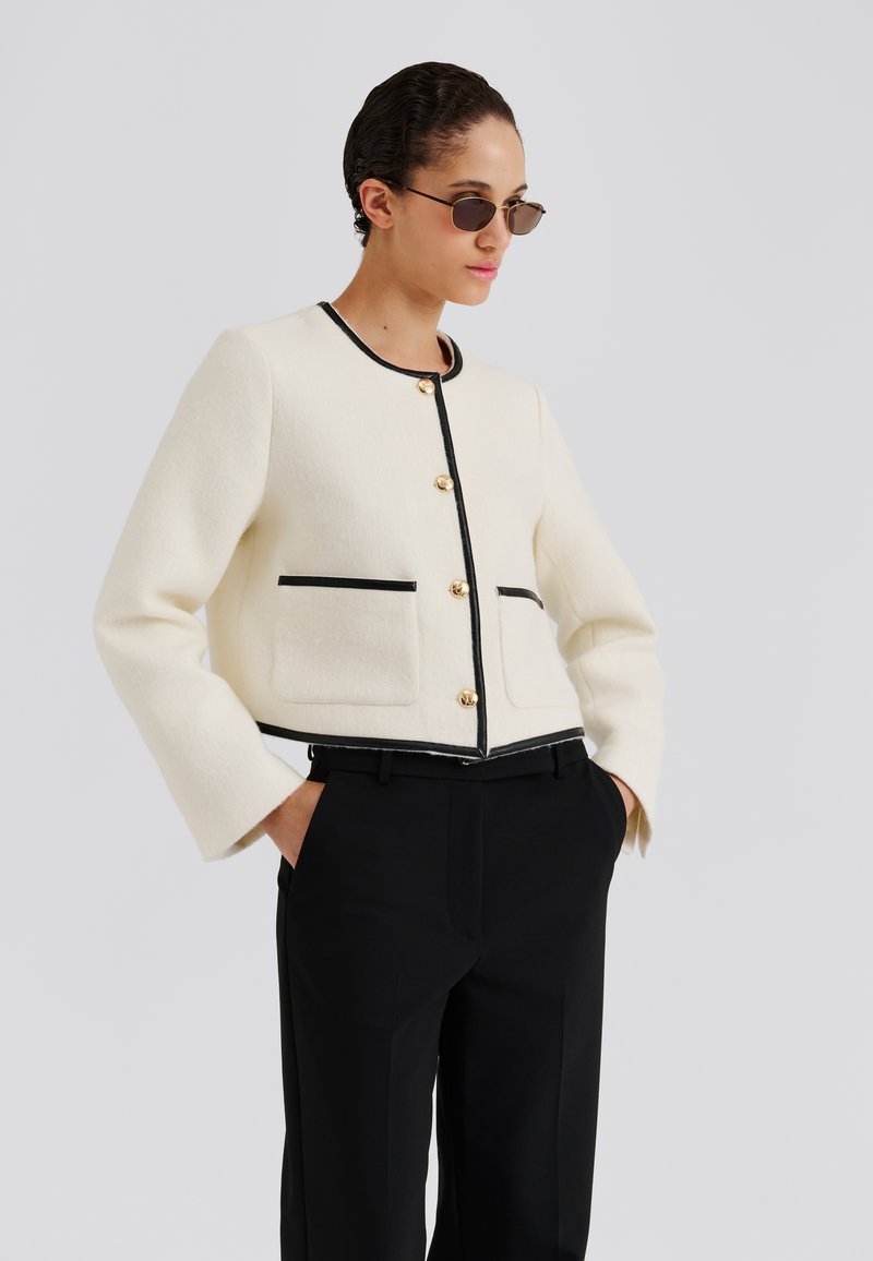 Biała kurtka z mieszanki wełny z czarnymi wykończeniami, złotymi guzikami i przednimi kieszeniami. Noszona z czarnymi spodniami, ma skrócony fason i rozszerzane rękawy.