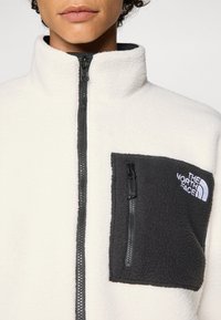 Biela fleecová bunda s vysokým golierom, predným zipsom a sivou náprsné kapsou so zipsom a logom "The North Face".