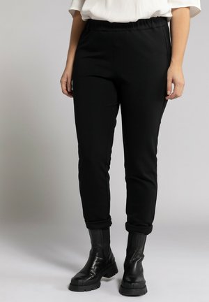 Pantalons de sport noirs avec une taille élastique, coupe ajustée et poignets retournés, associés à des bottines noires, sur un fond neutre.