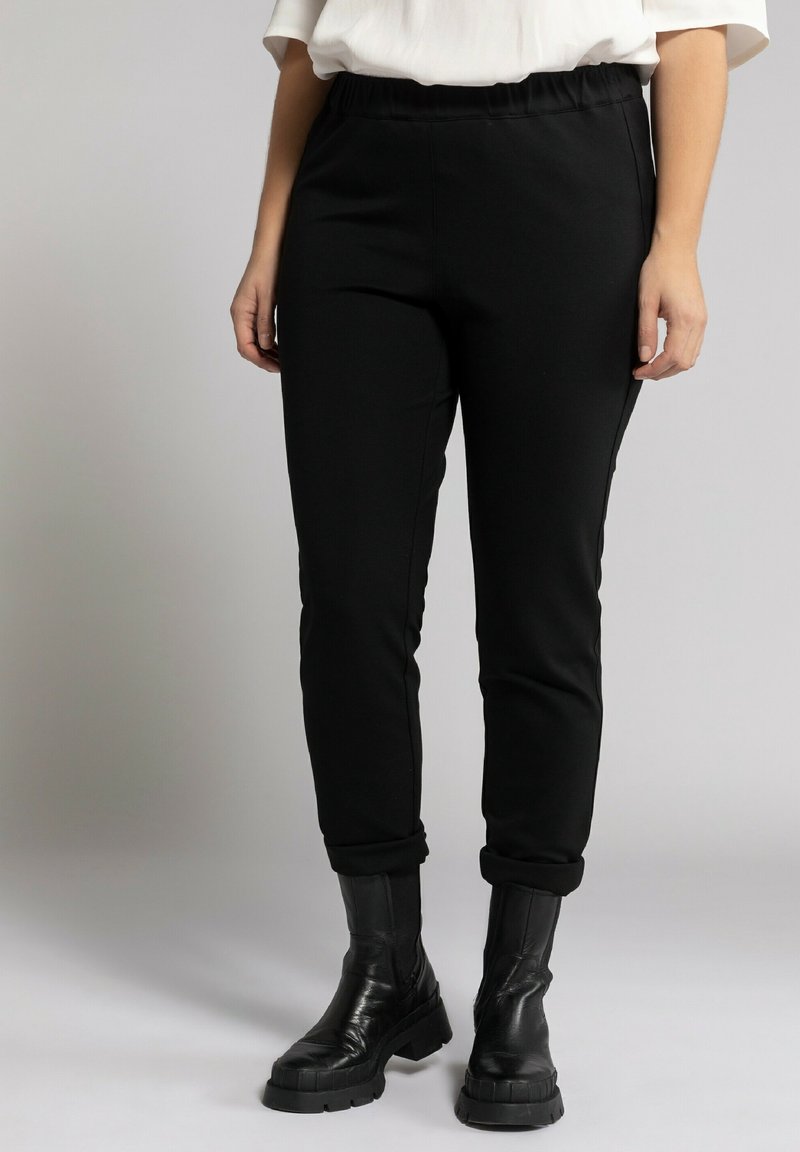 Pantalones de yoga negros con cintura elástica, corte ajustado y puños enrollados, combinados con botas negras hasta el tobillo, sobre un fondo neutro.
