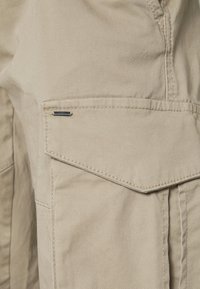 Beige stoffen cargobroekzak met flap en een klein metalen label bevestigd nabij de bovenrand.
