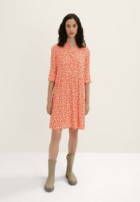 Robe orange avec un motif floral blanc, manches 3/4, décolleté en V et jupe évasée, assortie à des bottes grises claires jusqu'aux genoux.