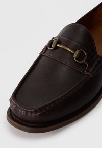 Mocasín de cuero marrón oscuro con detalle de estribo de latón y diseño de puntera cosida sobre fondo blanco.