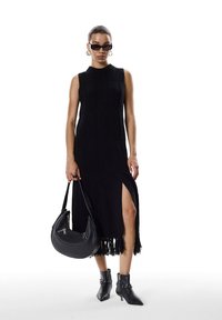 Robe sans manches en tricot noir avec fente sur le côté, ourlet à franges et texture côtelée. Associée à un sac à main noir et des bottines.