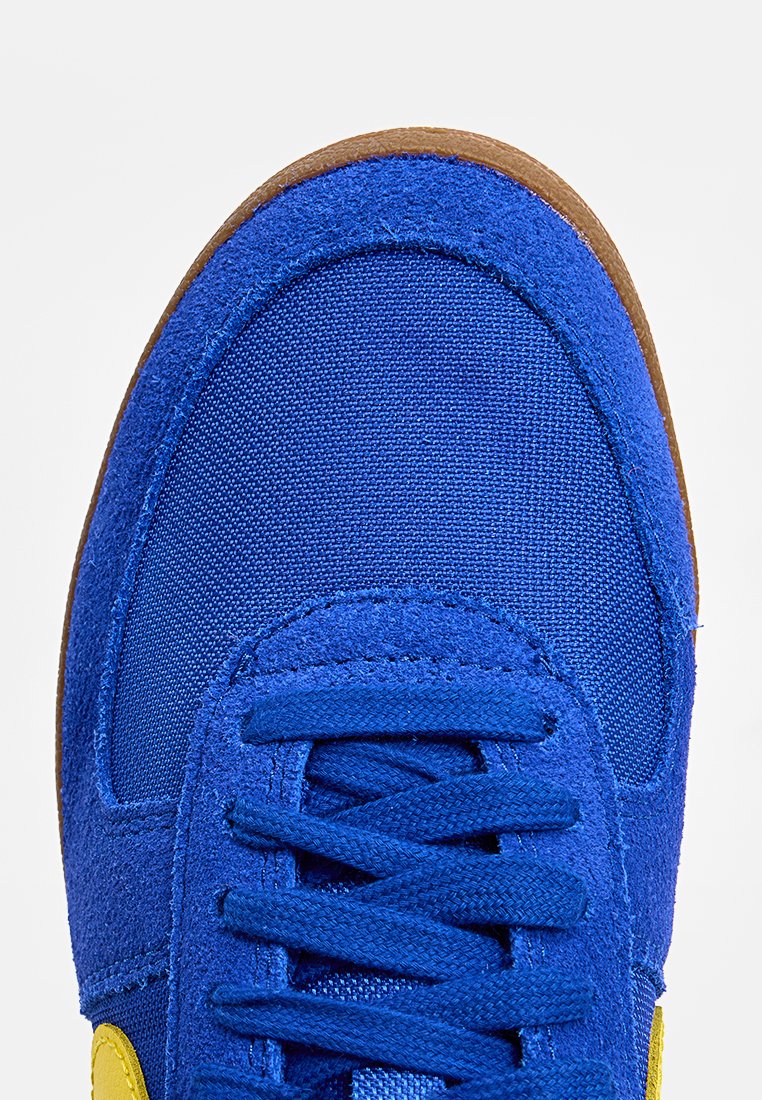 Blaue Sneaker mit einem Obermaterial aus Wildleder und Textil, runder Zehenpartie, Schnürdesign, Gummisohle und einem auffälligen gelben Akzentlogo.