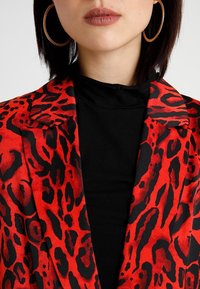 Punainen ja musta leopardikuosinen bleiseri, jossa on lovikaulus, puettu mustan topin päälle. Korostaa rohkeita kuvioita ja sileää kangaspintaa.