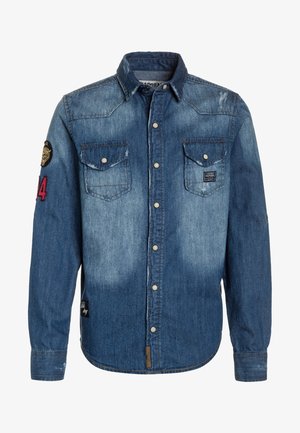 Koszula dżinsowa w różnych odcieniach niebieskiego, z wyraźnym kołnierzykiem, dwoma kieszeniami na piersi i zatrzaskami. Zawiera łatę na rękawie.