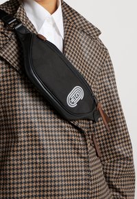 Černá látková křížová taška s texturovaným povrchem, která má zipovou kapsu a detail s logem, nošená přes hnědý kostkovaný kabát.