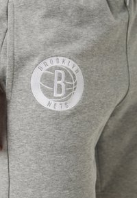Pilkos sportinės kelnės su baltu siuvinėtu emblemą, kurios viduje yra žodžiai "BROOKLYN" ir "NETS", supančios krepšinio dizainą.