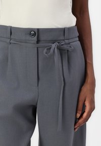 Pantalons tailleur gris avec une texture lisse, dotés d'une fermeture à bouton à l'avant, de passants de ceinture et d'une ceinture en tissu nouée.