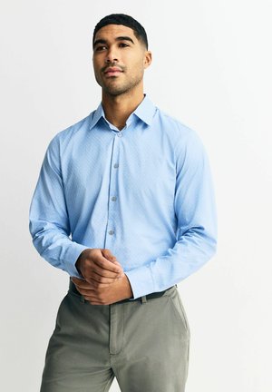 Mann mit kurzen Haaren, der ein hellblaues Hemd mit Knöpfen und graue Hosen trägt, steht mit verschränkten Händen und schaut leicht nach oben.