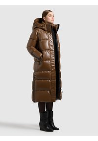 Ruda, paminkštinta ilga striukė su blizgiu paviršiumi, kapišonu ir šoniniais užtrauktukais. Dėvima ant juodo megztinio, derinama su juodais kulkšnių ilgio batais.