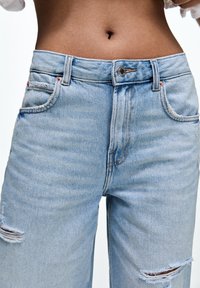 Midja på person som bär ljusblå, slitna denimjeans med knäppning och blixtlås, där bar hud och navel syns.