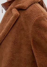 Primer plano de un abrigo teddy marrón hecho de una tela suave y texturizada, con amplias solapas y un vistazo a un cuello de cuero marrón debajo.