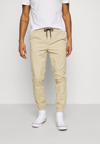 Beige, lightweight jogger pants with an elastic waistband and black drawstring, featuring elastic cuffs and a smooth texture. Worn with white sneakers.