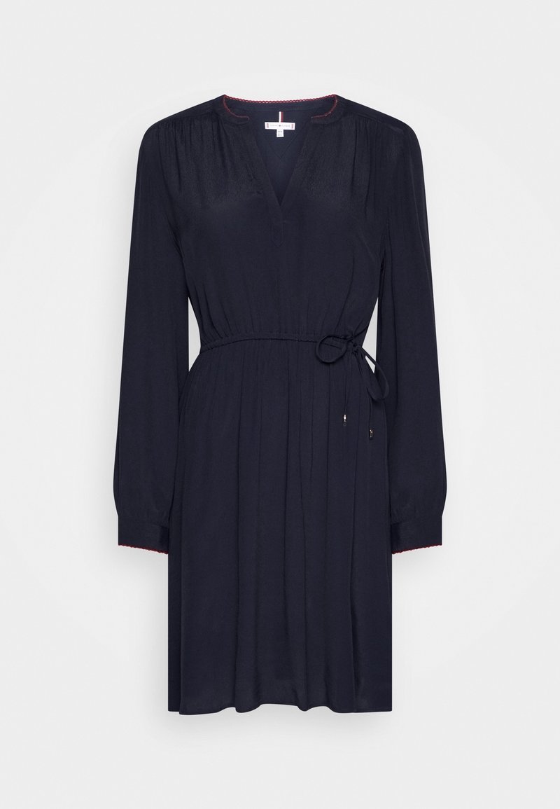 Robe noire à manches longues avec col en V, taille froncée nouée avec une fine ceinture, et détail subtil de surpiqûres rouges sur le col et les poignets.