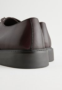 Tmavoburgundské kožené topánky s hladkým povrchom, okrúhlym špičkou a hrubou čiernou gumovou podrážkou. Viditeľné prešitie pozdĺž švov.