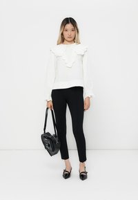 Giovane donna che indossa una blusa bianca con volant e pantaloni neri aderenti, mentre tiene una borsa a forma di cuore nera, in piedi su uno sfondo bianco.