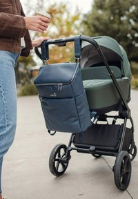 Tmavě modrá texturovaná taška na plenky se zipy připevněná k elegantnímu kočárku. Kočárek má zelený stříšku a černá kola na šedém povrchu.