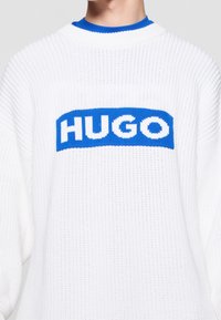 Biały dziany sweter o luźnym kroju. Posiada niebieską prostokątną naszywkę z napisem "HUGO" w pogrubionych białych literach.