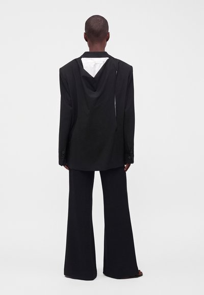 Personne aux cheveux courts portant un blazer noir oversize avec capuche et un pantalon noir à jambes larges, debout face à l'arrière sur un fond uni.