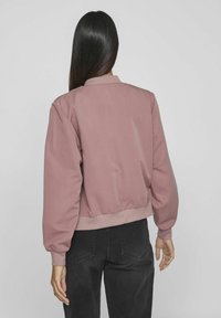 Woman with long straight hair wearing a dusty pink bomber jacket and black jeans, standing facing away against a plain gray background.