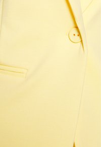 Žlutý blazer vyrobený z hladké tkaniny, s jedním žlutým knoflíkem, malou kapsou a přiléhavým střihem.
