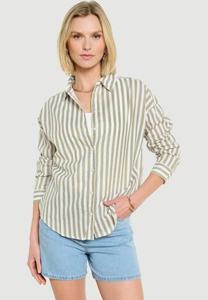 Blondínka v olivovo-bielej košeli s vertikálnymi prúžkami a svetlomodrých džínsových šortkách, so zlatým náhrdelníkom a náramkami, ruka v vrecku.