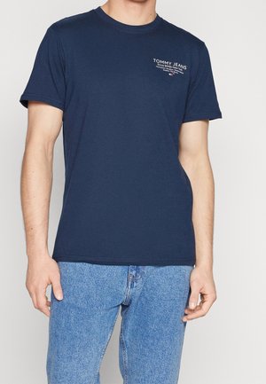 Homme portant un t-shirt bleu marine Tommy Jeans avec un logo blanc sur la poitrine gauche et un jean en denim bleu clair.