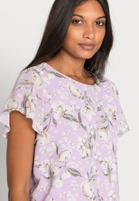 Top floral roxo com mangas curtas e franzidas. Apresenta um padrão de flores em tom creme e folhas verdes numa tecido liso e leve.
