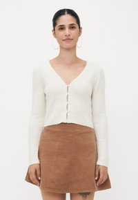 Valkoinen ribbed neulecardigani, jossa on V-pääntie ja viisi nappia, yhdistettynä ruskeaan corduroy-minihameeseen, jossa on viistoja saumoja.