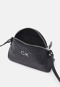 Borsa a tracolla in ecopelle nera con logo 'CK' impresso, hardware argentato, chiusura con zip e tracolla regolabile. L'interno è foderato in tessuto nero.