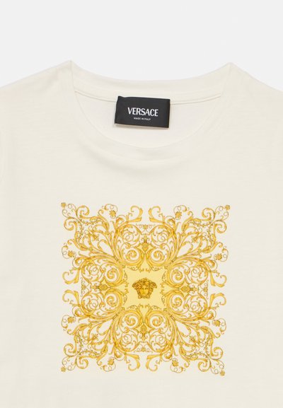 Λευκό T-shirt με χρυσαφένιο γραφικό σε μπαρόκ στιλ, που απεικονίζει περίτεχνες σπείρες και μια κεντρική κεφαλή Μέδουσας, συνοδευόμενο από μια μαύρη ετικέτα Versace.